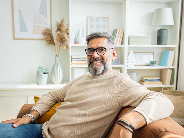 Smiling middle aged white man with glasses and a beard sits on a couch in a living room