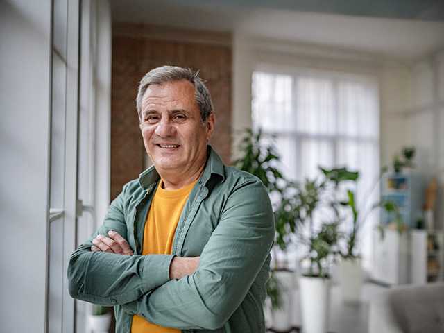 Grey-haired white man wearing a yellow shirt and green jacket crosses his arms while standing in a sunny room
