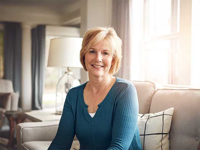 White woman in a teal sweater smiles while she sits in a sun filled room