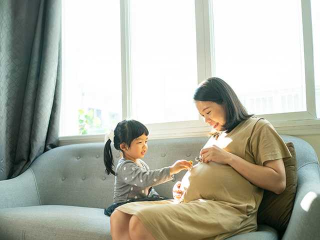 Pregnant Asian woman sits on a couch as her young daughter touches her belly