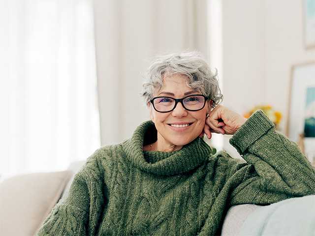 Smiling older white woman with short grey hair sits on a couch in a sunlit room