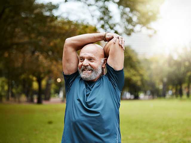 White man with a bald head and grey beard stretches his arms over his head while standing outdoors