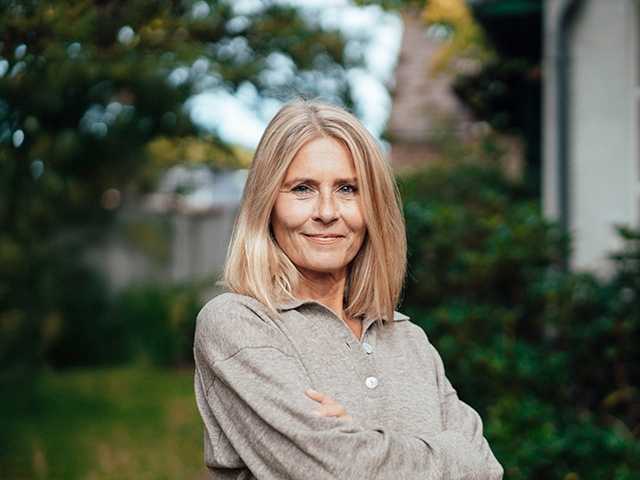 White woman wearing a tan sweater crosses her arms as she stands outside in front of trees