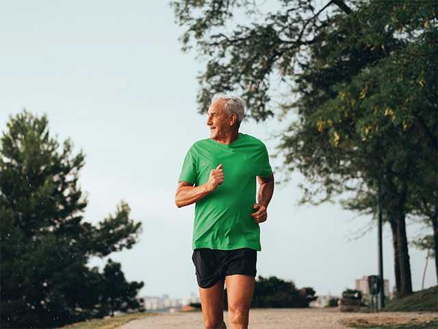 Older man wearing a green shirt runs down a paved path outdoors in the sunset