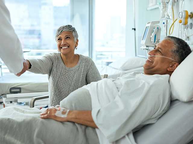 Male patient in a hospital bed watching while his female visitor smiles and shakes hands with the patients doctor