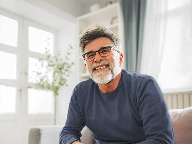 Older white man with a grey beard and glasses sits in a sunny room