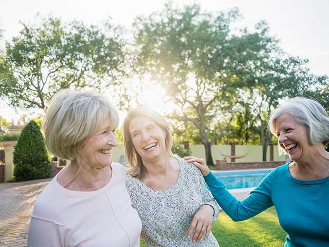 Three older women smile and laugh as they stand together in the evening sunshine
