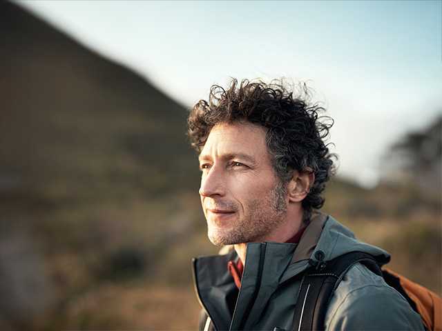 Middle aged white man with curly brown and grey hair stands in the evening sun with a mountain in the background