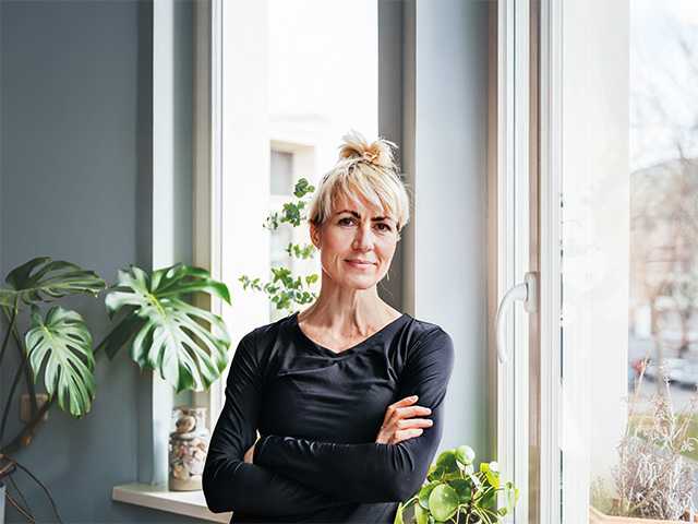 Middle aged white woman wearing a black shirt stands in a sunny room, crossing her arms over her torso