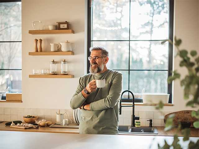 Smiling man with grey hair and a grey beard stands in a kitchen holding a coffee mug