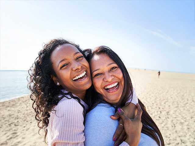 Smiling young woman with curly brown hair hugs her smiling mother while they stand in the sunshine on a beach