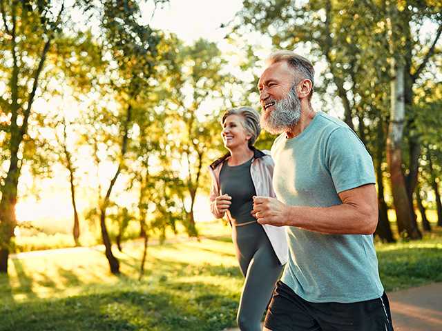 Man with grey hair and a grey beard runs through a sunny wooded area alongside a woman with grey hair