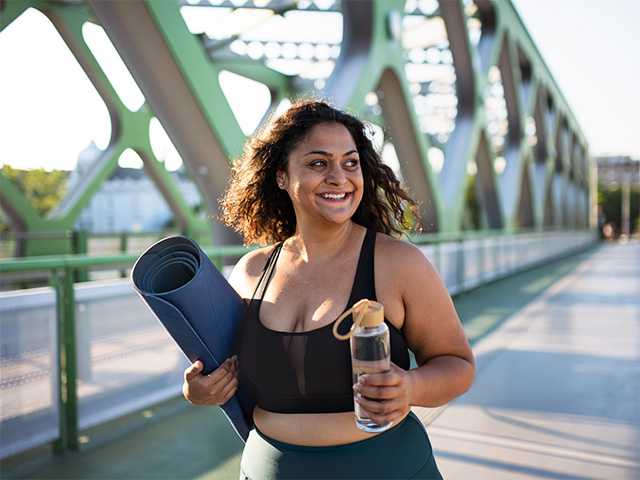 Middle aged woman carries a yoga mat and a water bottle outdoors in the sunshine