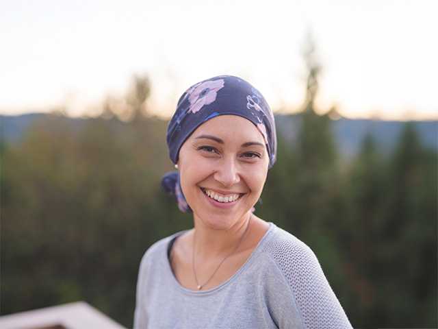 Smiling young white woman wearing a purple bandana on her head stands outdoors in the evening sunlight