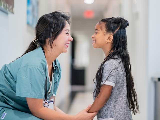 Smiling woman in light blue scrubs holds hands with young girl in hospital hallway