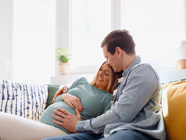 Young man hugs his pregnant wife and places a hand on her belly as they sit on a couch