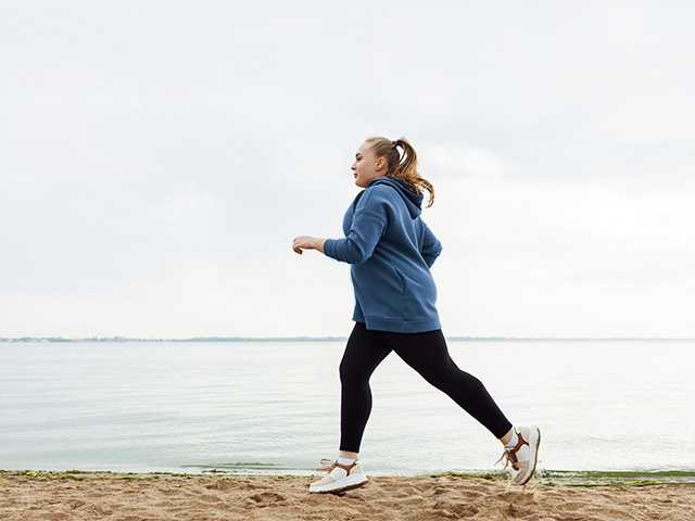 Middle aged white woman in long pants and a sweatshirt jogs alongside a lake