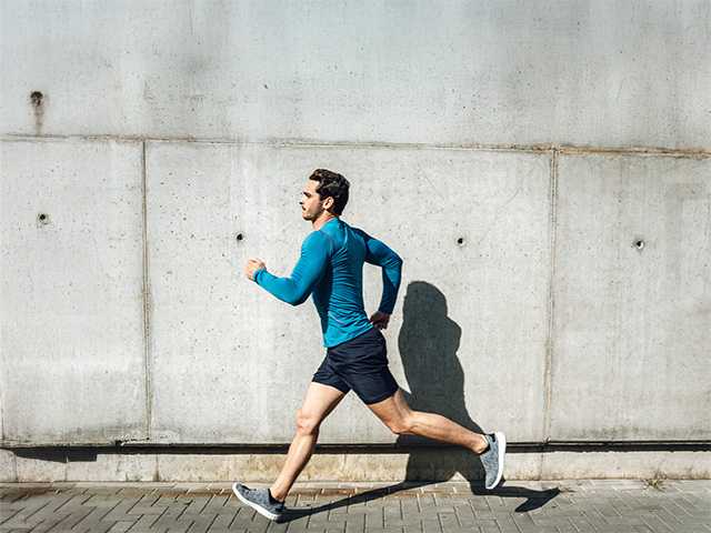 Young white man in black shorts and a blue shirt jogs on the sidewalk in sunshine