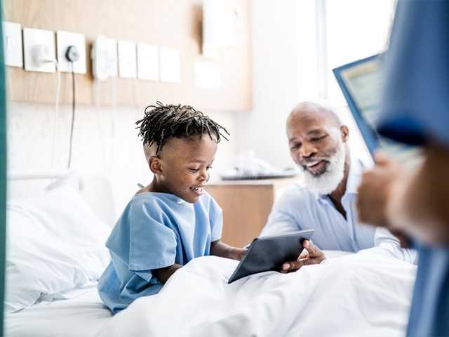 Smiling young Black boy in a hospital bed looks at a tablet with his father next to him