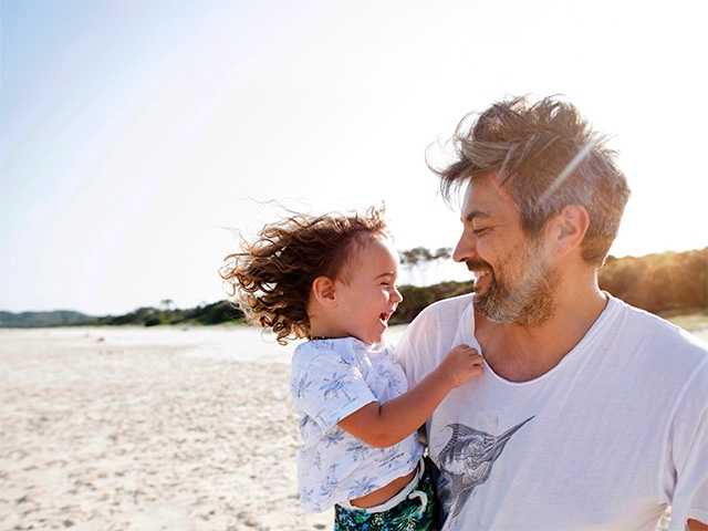 Middle-aged white man holds his toddler son while standing in the sunshine on the beach