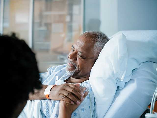 Older Black man laying in a hospital bed smiles and holds hands with his wife