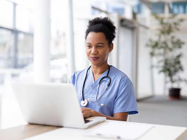A woman in scrubs sits at a desk, focused on her laptop, surrounded by medical supplies and paperwork.