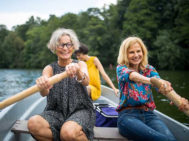 Two smiling white women sit side-by-side rowing a boat in a lake