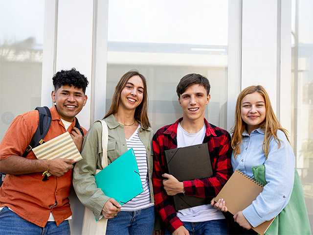 Four students stand together, smiling, each holding a stack of books in a bright, outdoor setting.
