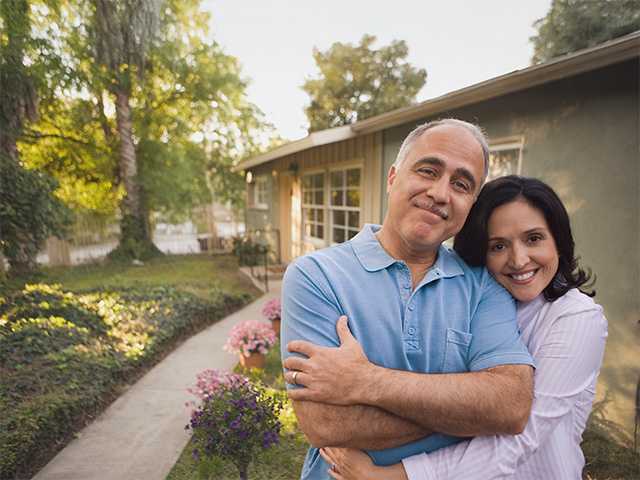 Man and his wife stand next to one another outdoors in the evening next to their home