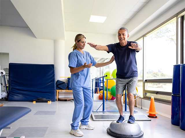 Woman in light blue scrubs helps a man as he balances on equipment in a rehabilitation gym