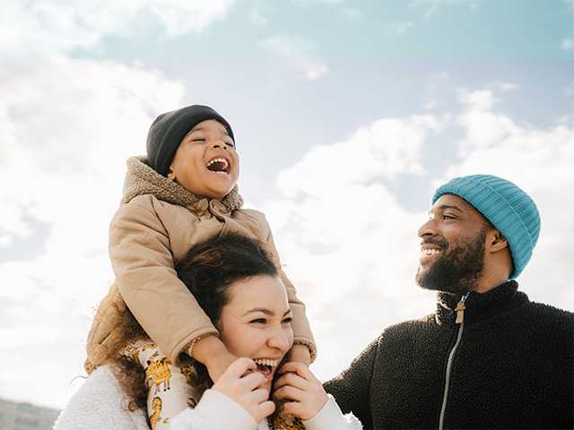 Smiling mother and father walk outdoors in the sunshine, as their young son laughs and sits on his mother's shoulders