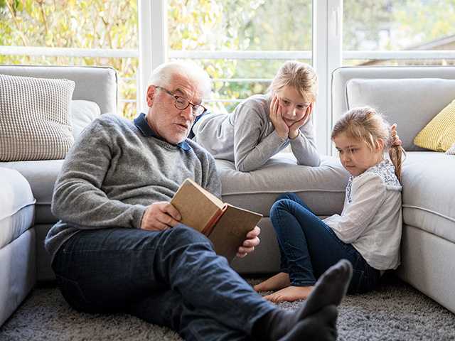Elderly white man sits on the floor and reads a book to his two granddaughters