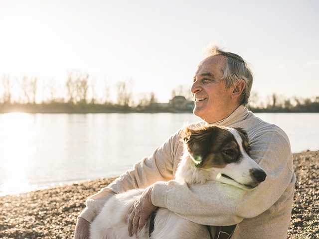Older white man wearing a white sweater sits with a dog next to a small lake