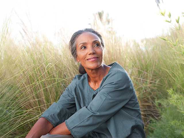 Black woman in a blue shirt sits in long wild grasses in the sunshine