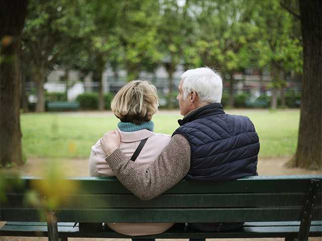 Elderly man sits next to his wife on a park bench, with a hand on her shoulder