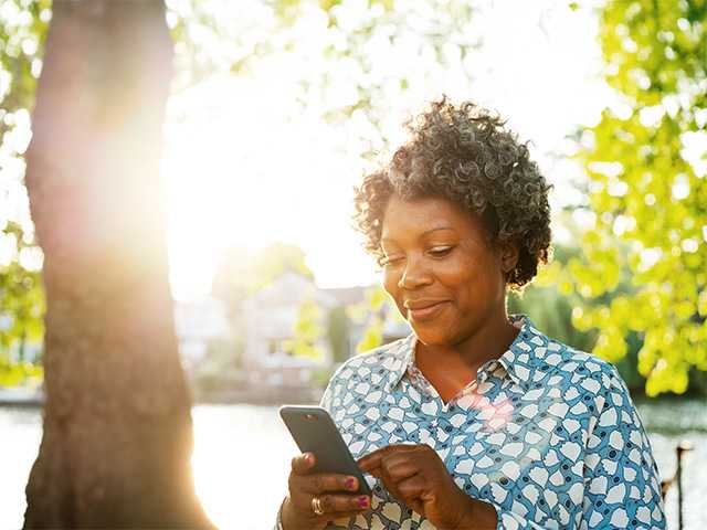 Black woman in a light blue shirt stands beneath a tree in the sunlight, looking at her phone