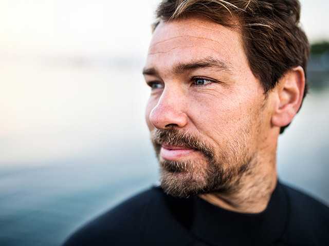 Man with brown hair and a goatee stands next to a lake, looking into the distance