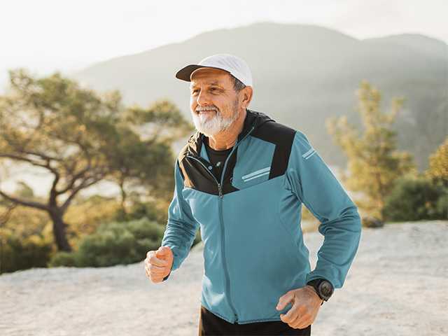 Older man with a grey beard runs outdoors while wearing a light blue jacket