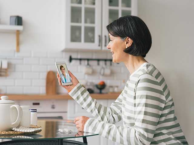 Woman with short black and grey hair sits in a kitchen speaking on a video call on her phone