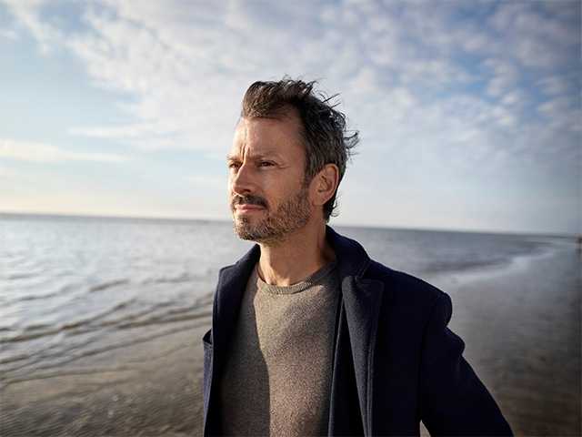 Middle aged white man wearing a navy blue zip-up sweatshirt stands on a beach