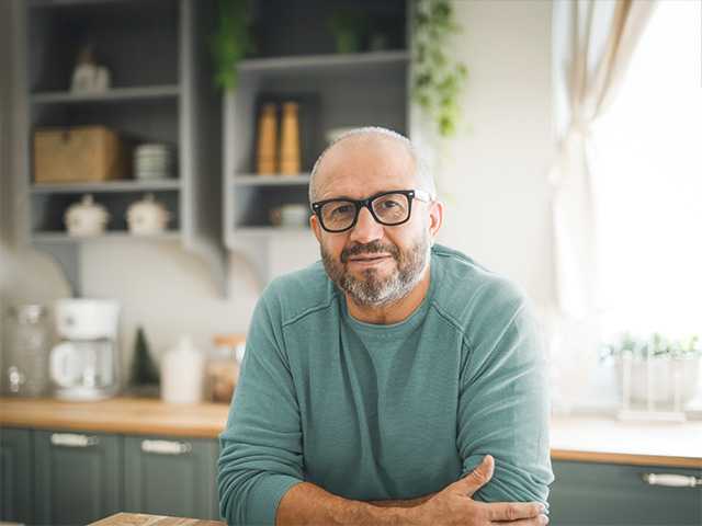 Older white man wearing glasses and a light blue shirt leans on the countertop in the kitchen