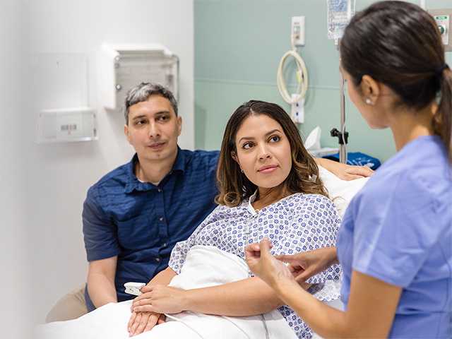 Woman in a hospital gown sits in a hospital bed, next to her husband, as they speak with a Corewell Health blood and marrow transplant specialist