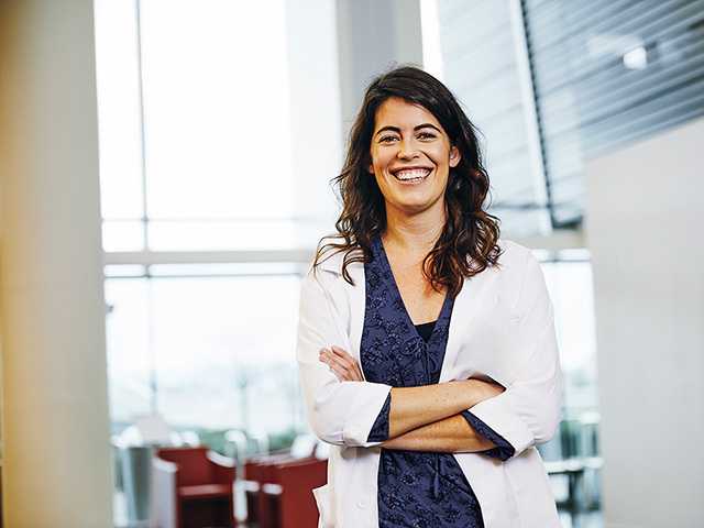 A female health care professional in a white coat is positioned in a hallway of an academic environment.