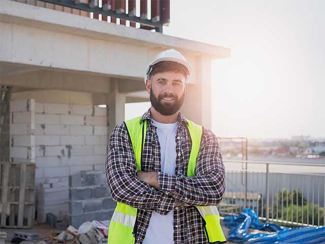 Smiling white man wearing a neon yellow safety vest and construction helmet stands on a work site in the morning sun