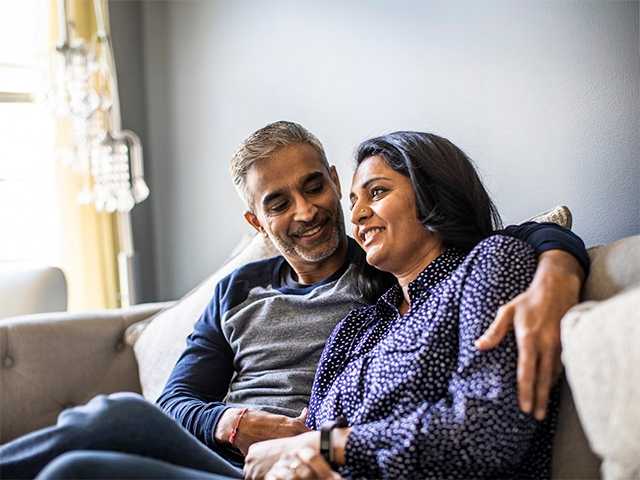 Smiling middle aged Indian man and woman sit beside one another on a couch hugging