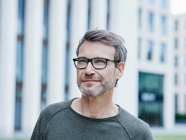 Middle aged white man with glasses and a grey beard stands outdoors in front of an office building