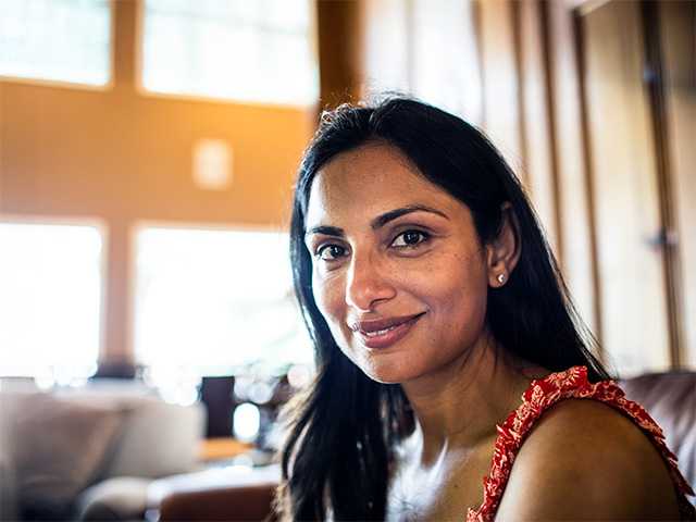 Smiling woman with long black hair sits indoors near a sunshine filled window