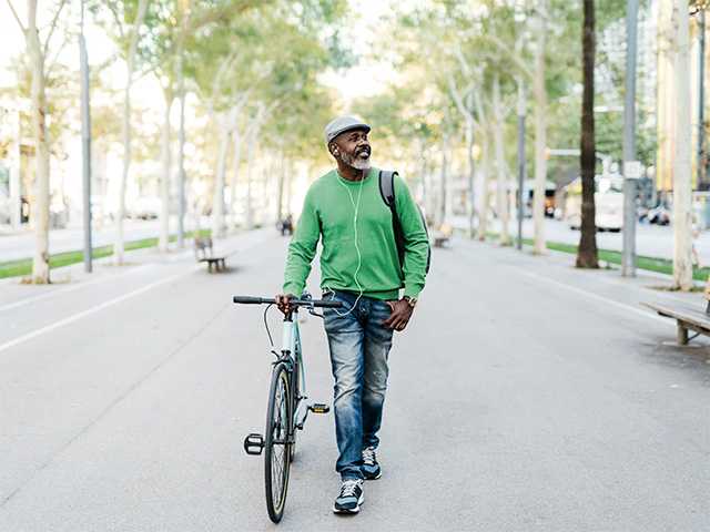 Middle aged Black man wearing a green sweater walks his bike through a park