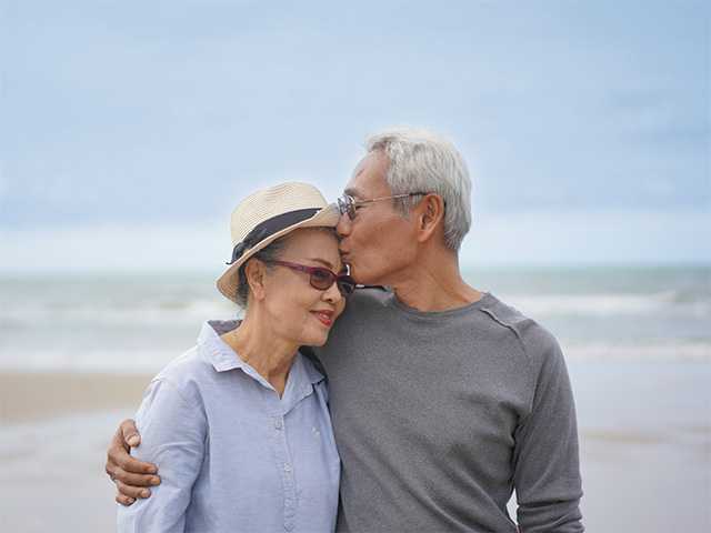 Older Asian man with grey hair and glasses hugs and kisses his wife