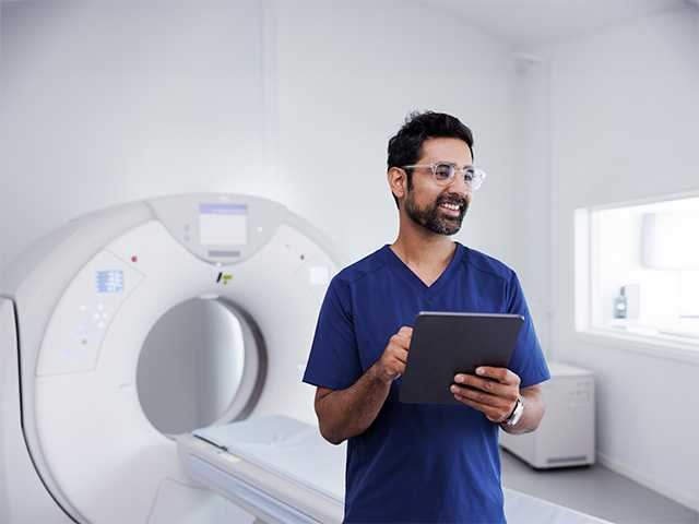 Man with glasses and a beard and wearing blue scrubs stands in front of an MRI machine